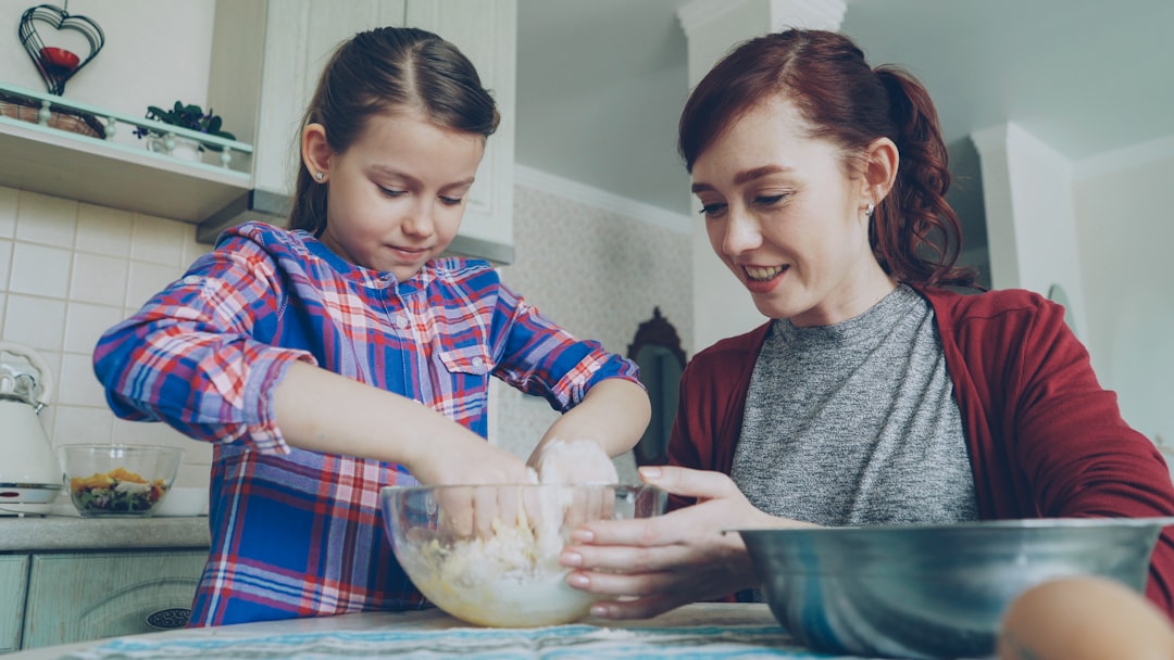 Little pretty girl helping her mother in the kitchen stirring dough for cookies into bowl. Mom and daughter have fun talking and laughing. Family, food, home and people concept
