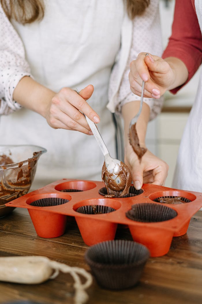 Two people preparing chocolate cupcakes with silicone molds on a wooden table.